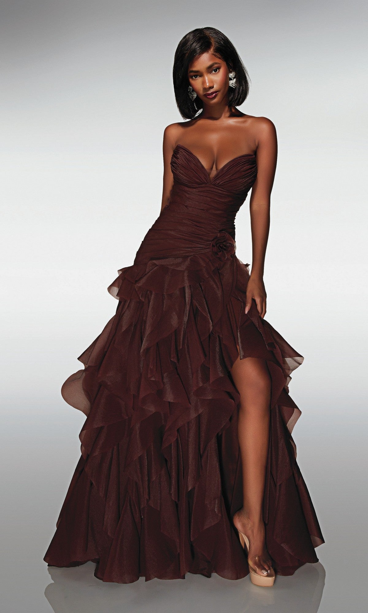 A woman poses in the strapless chocolate Alyce 62024 long prom dress with ruffles and a high slit, standing against a plain gradient background.
